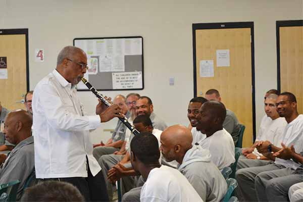 A male student plays a clarinet in front of a group of other students. Everyone is wearing a gray uniform and appear to be sitting in an institution.