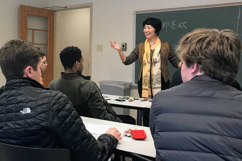 Chinese Instruction An instructor smiles while talking at the front of a classroom, as students are seen from behind. Chinese characters are visible on the board in the background.