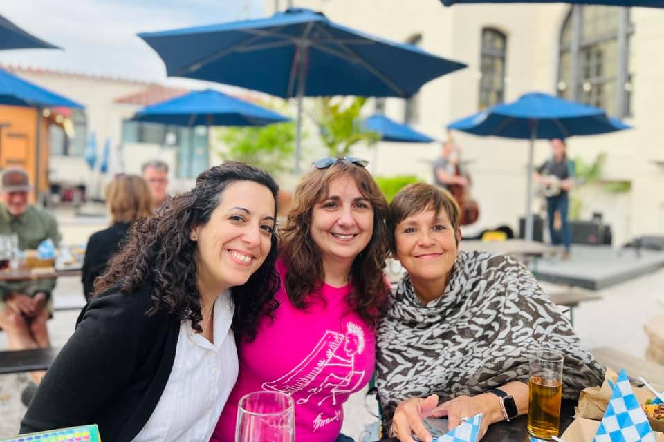 Slu graduate student gathering Three students sit next to each other and smile at the camera at an outdoor gathering.