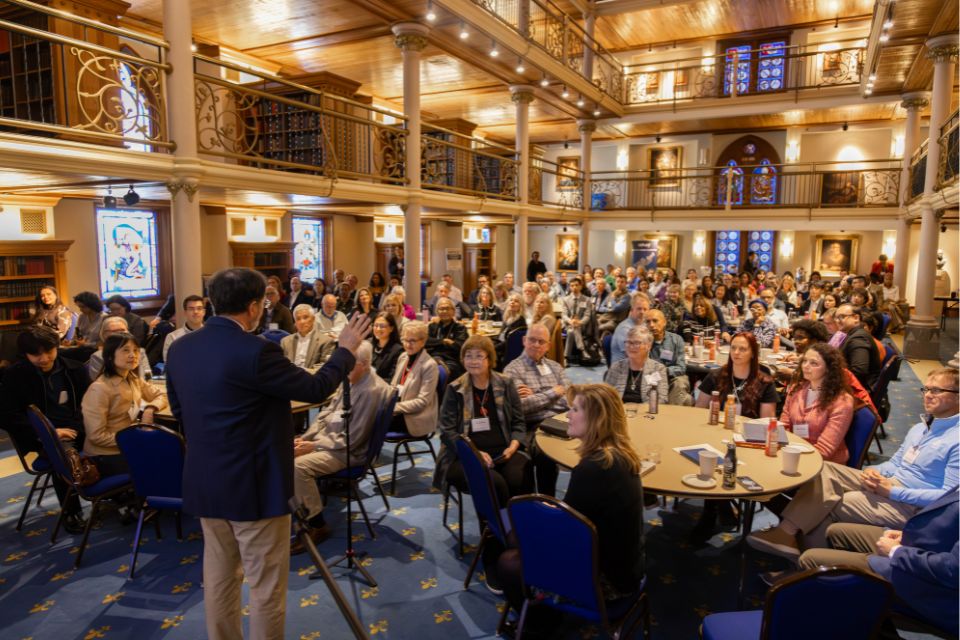 Leadership Speaker Series Audience A crowded room of conference attendees are seen from the front while sitting at tables, watching a speaker