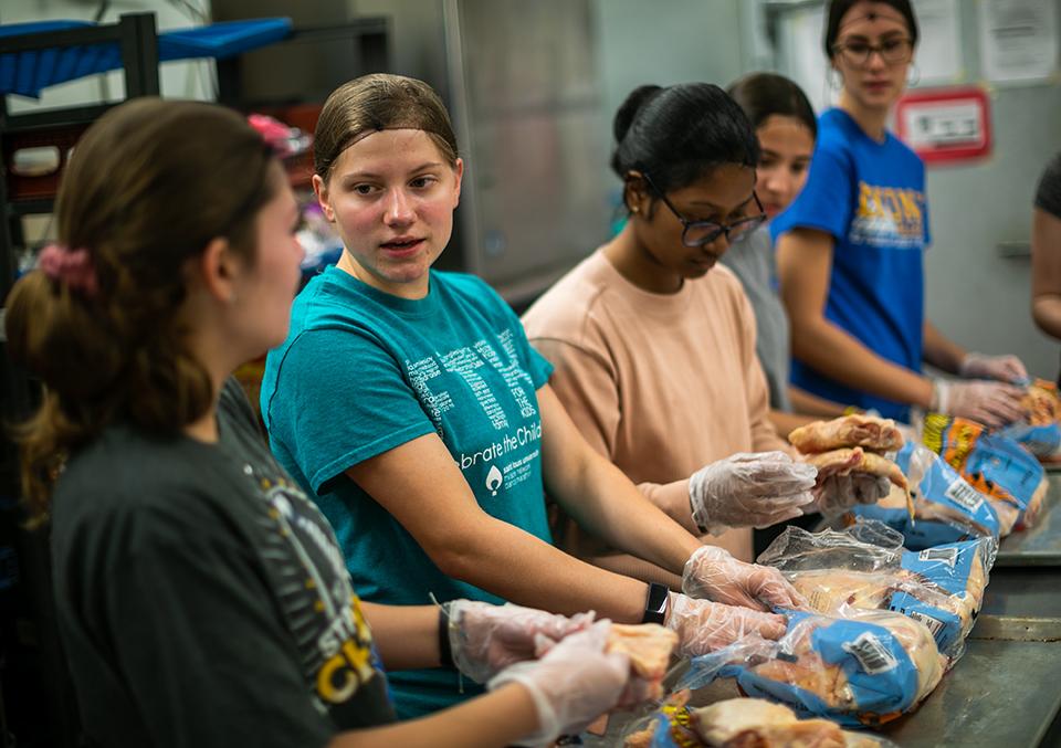 Students working in Campus Kitchen Students wearing hairnets and gloves work in a line at a kitchen counter.