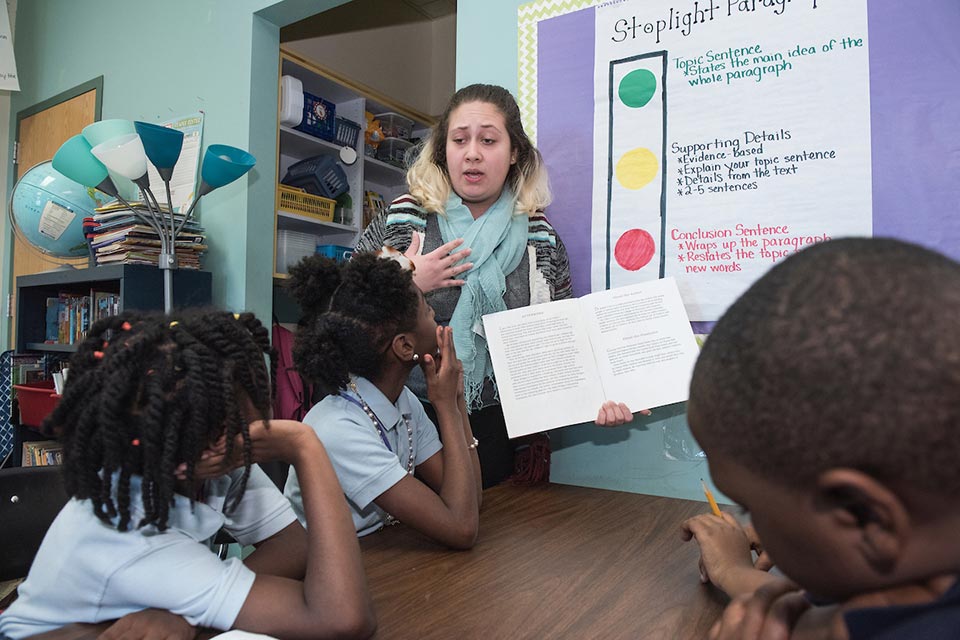 Academic center A teacher holds a book open while elementary students sit at a table in a classroom. A bulletin board behind the teacher reads "stoplight paragraphs."