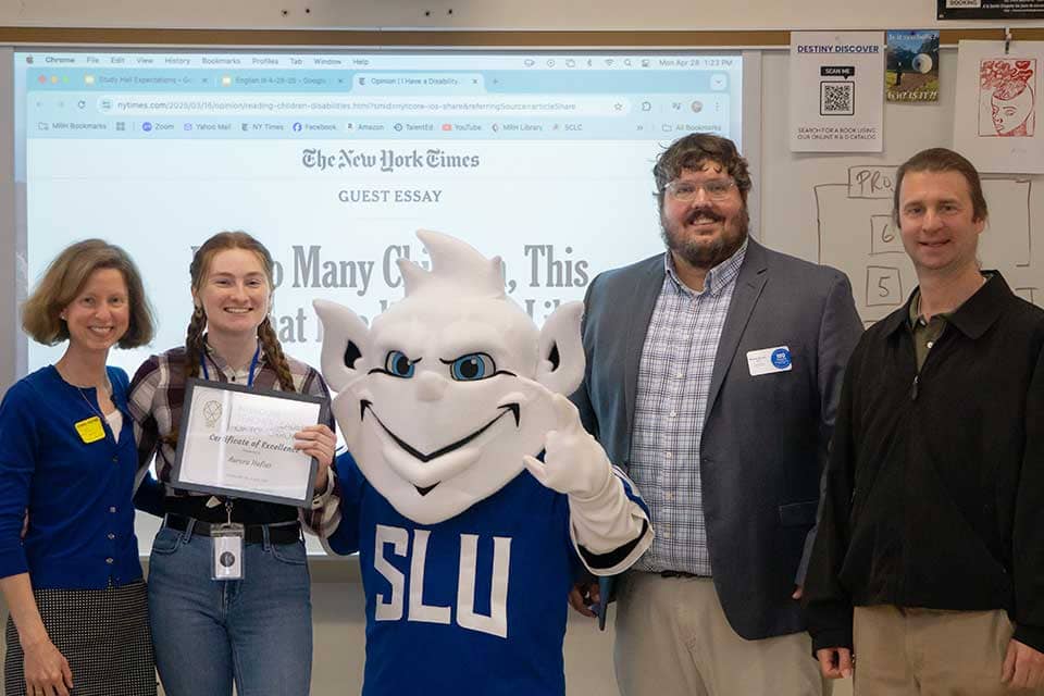 Hofius award photo Aurora Hofius, a young female student, stands surrounded by educators and the Billiken in front of a white board.