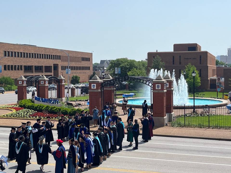 SOE pre commencement 2025 A group of faculty in academic regalia form a line and clap while graduates in caps and gowns cross the street. A bagpiper plays in the background.