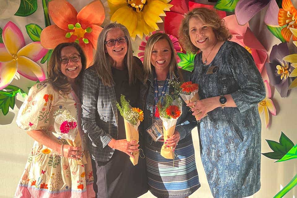 SLU Women Educators Celebration Four women pose in front of a wall covered in flower sculptures. Each one holds a small bouquet.