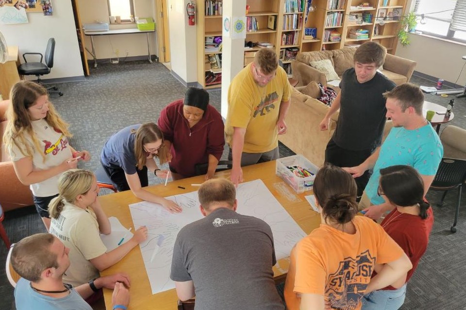 BTC Community Program participants stand around a table and examine a large piece of paper placed in the middle.