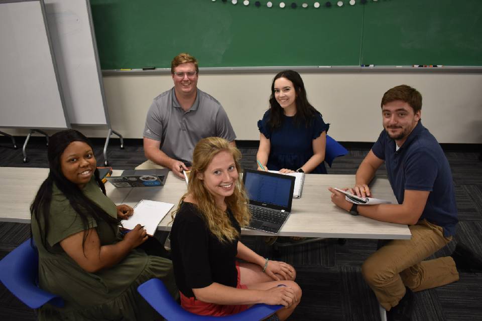 BTC MAT students in class BTC MAT look up and smile at the camera while sittng around a table with laptops and notebooks.