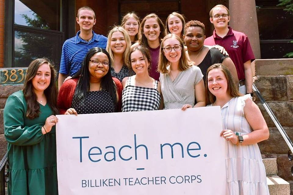 B T C Sign A group of 12 teachers corps members stand on steps outdoors while holding a sign that reads "Teach me. Billiken Teachers Corps."