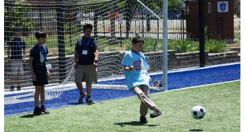 Access Academies 2024 Summer Program A student kicks a soccer ball on a field while two others, standing in a goal, watch.