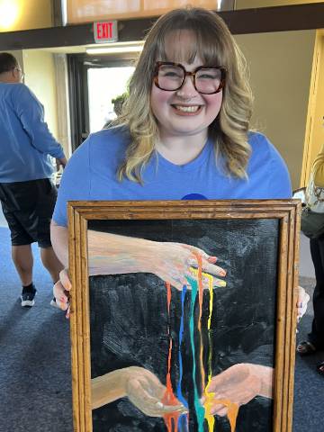 Anjelikka Hopkins with her painting A smiling woman holds a framed painting, which depicts a hand pouring streams of multiple colors of paint onto two hands below.