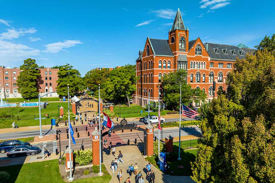 A view of campus from above. A tall brick building is seen and many people cross the street from one side of campus to another.