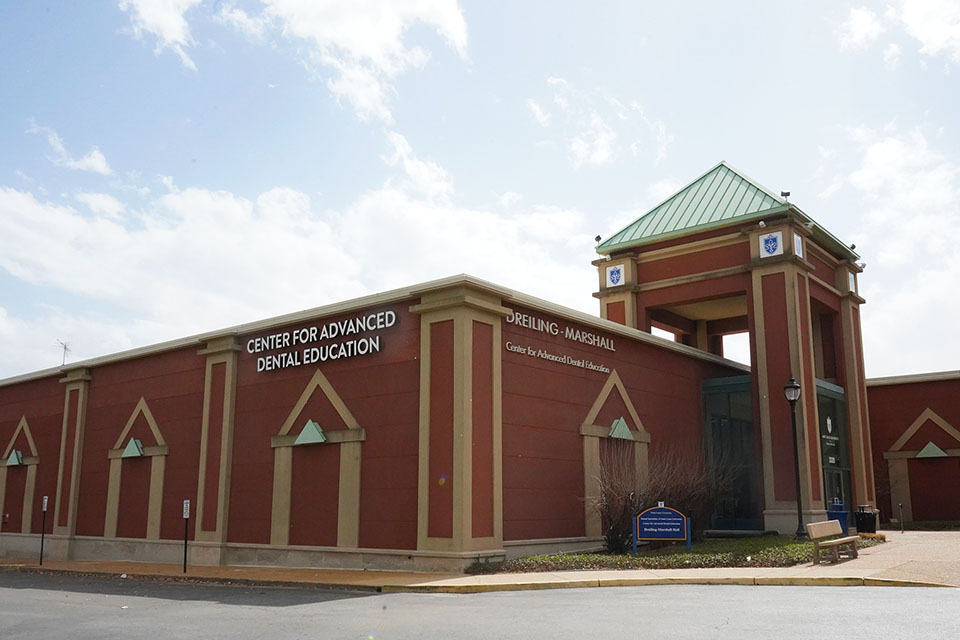Red-brick building with green roof, labeled "Center for Advanced Dental Education" with a hazy and cloudy sky backdrop.