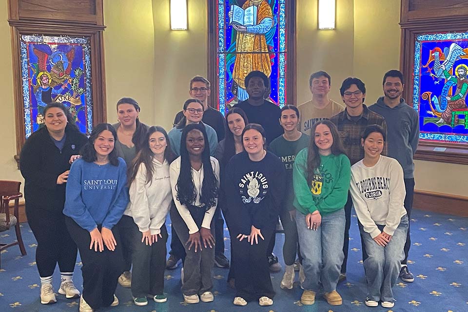 BEC student callers A group of 15 SLU students and their staff supervisor pose for a photo in a room at DuBourg Hall with stained glass windows behind them.
