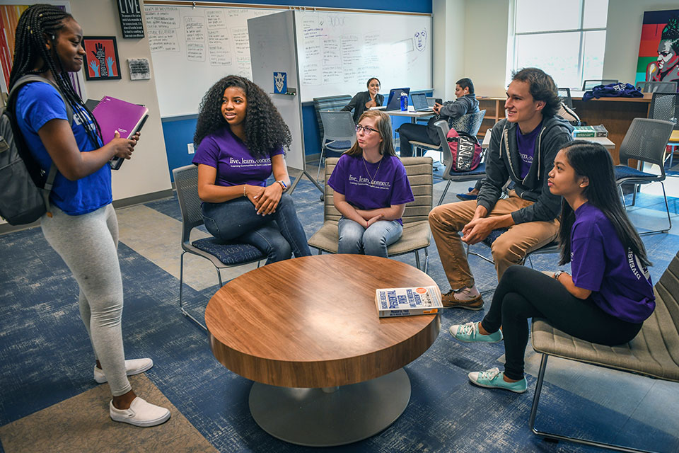 Diversity and Global Citizenship Four students, each wearing t-shirts that read "live. learn. connect." sit around a round coffee table while looking up at another student wearing a SLU Billikens T-shirt.