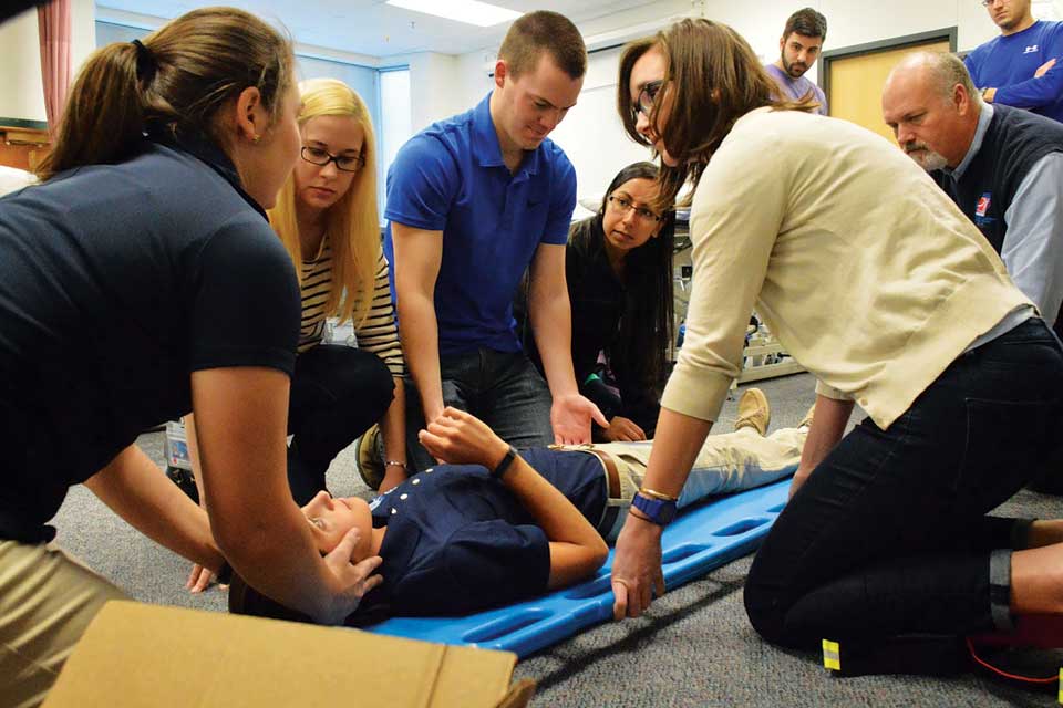 Health Sciences Students A student lies on a back board surrounded by other students and instructors during a demonstration.