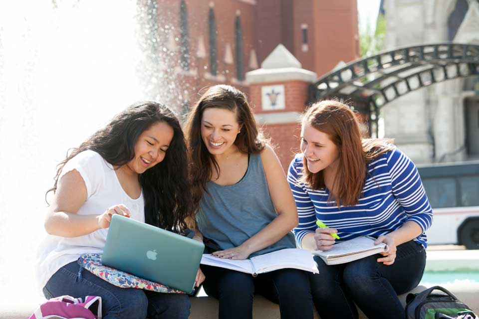 Honors Program Three students share a look at a laptop, with notebooks also on their laps, while sitting in front of a fountain.