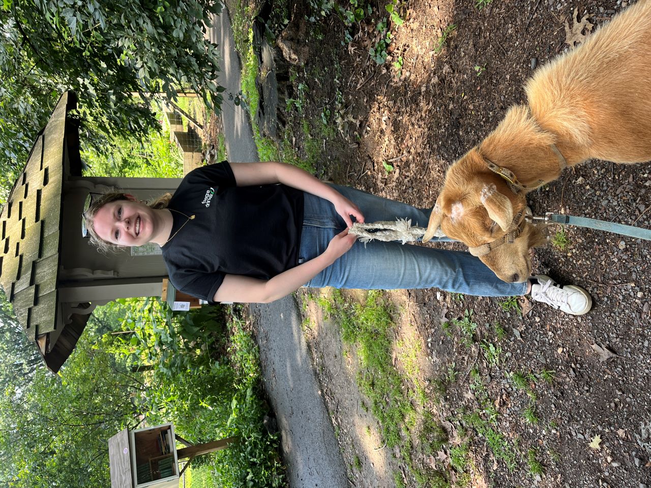 Katherine Tanis stands outside on a wooded walking path with a tan goat Katherine Tanis stands outside on a wooded walking path with a tan goat