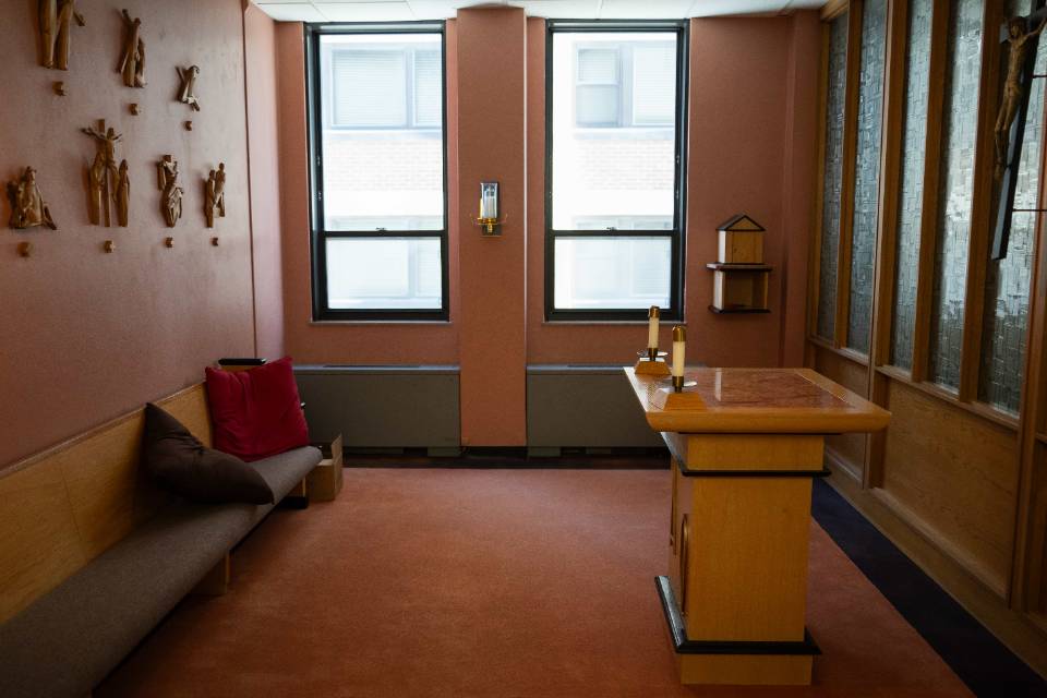 Demattias Hall Interior view of a room with an altar table on one side and a church pew on the other, with two tall windows on the end. Wooden depictions of the stations of the cross are on one wall and a crucifix is seen on the other.