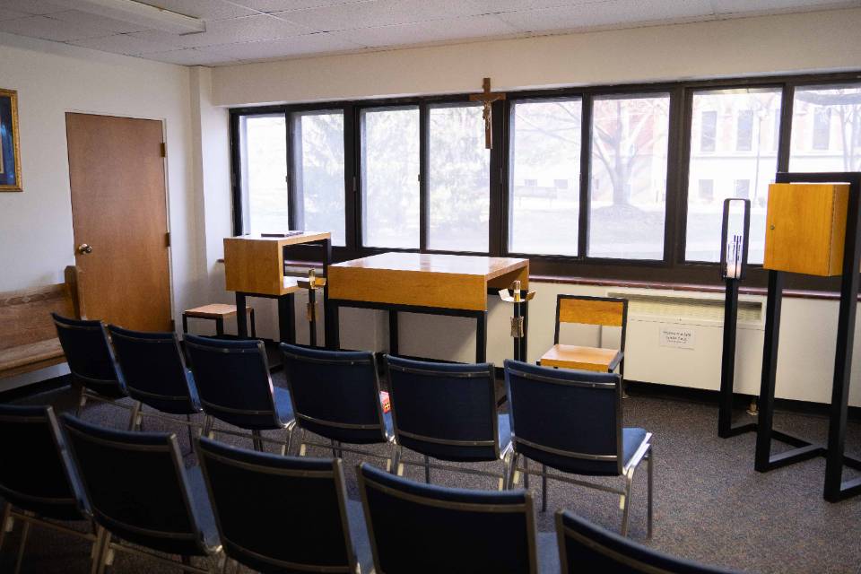 Fusz Hall Chairs are seen from behind in two rows, facing a wooden altar table, chairs, crucifix and candle holder, along with a bank of windows.
