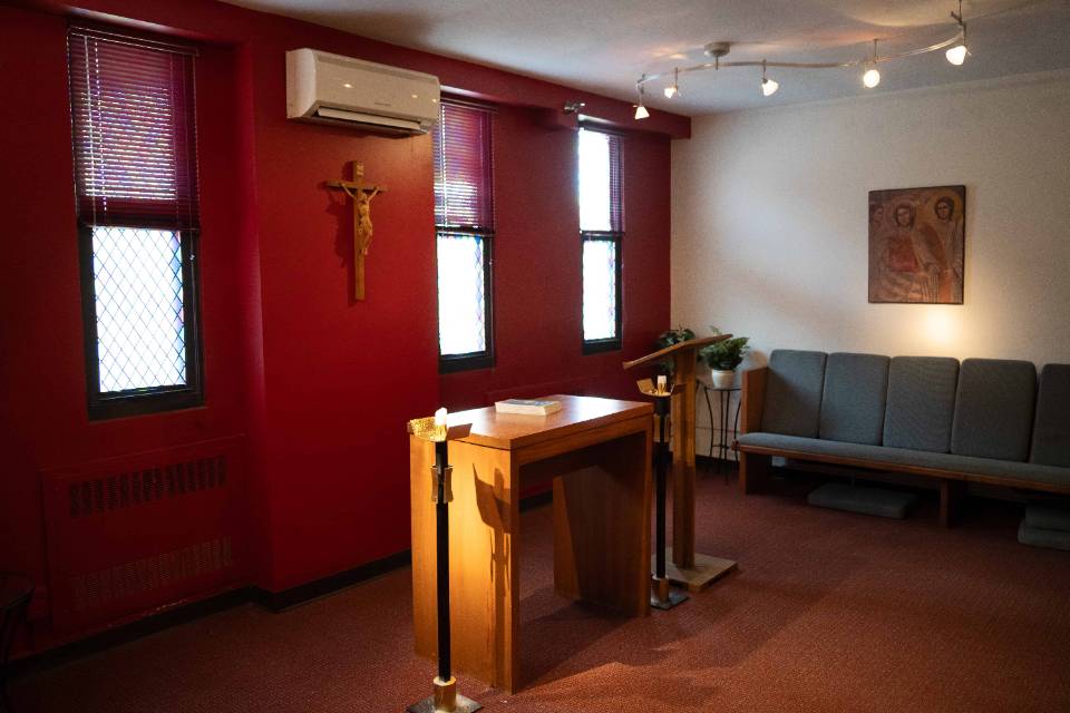 Greisedieck chapel Interior view of a room with an altar, crucifix lectern and candle holder at the front and center, with a padded pew along one wall. A religious painting is visible.
