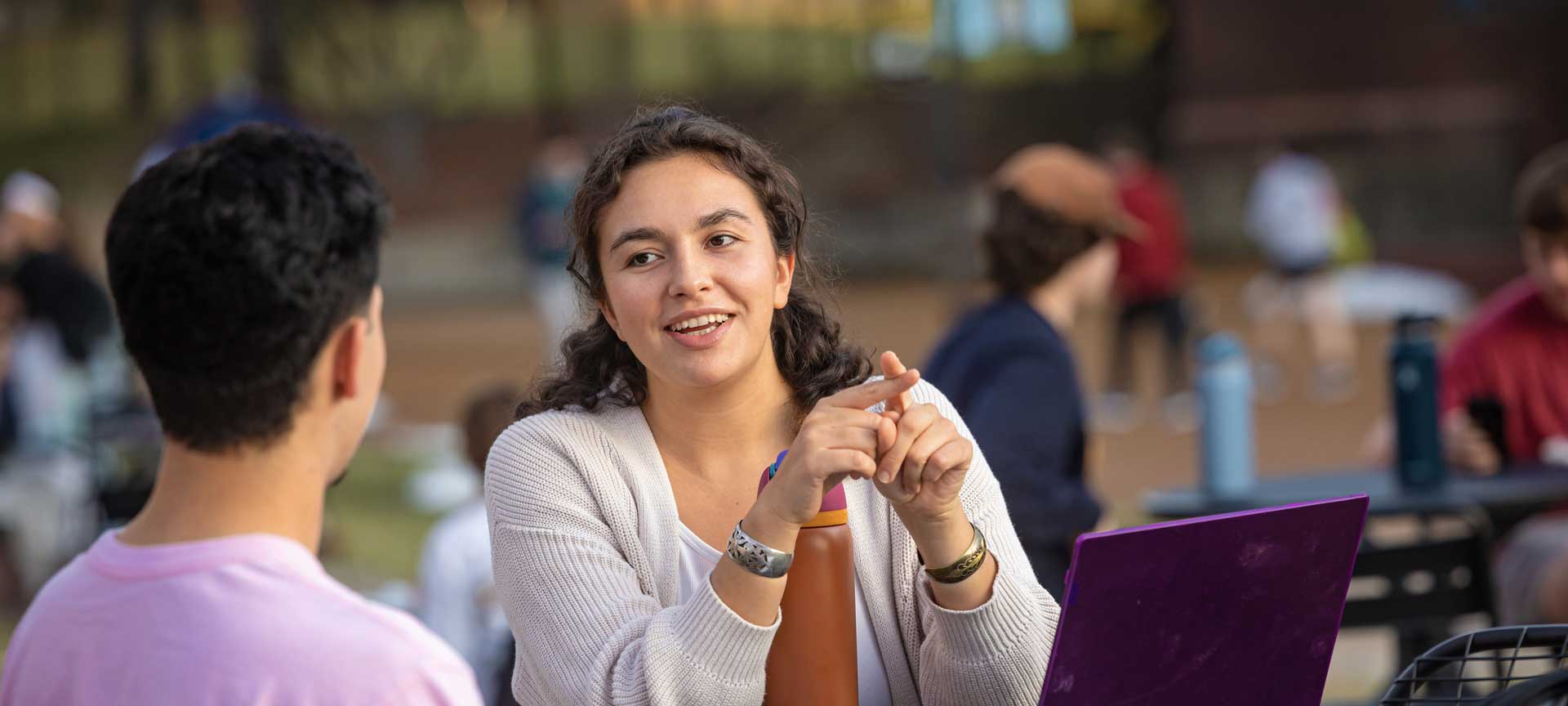 A student sitting at a table outdoors works on a laptop while conversing with another student.