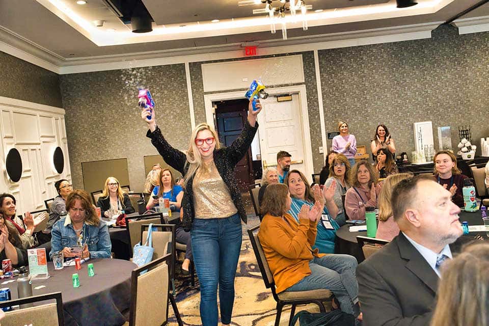 Erika Michalski Erika Michalski, a blond woman wearing red framed glasses, holds up two bubble makers while walking past event attendees who are seated at round tables in a large event space.