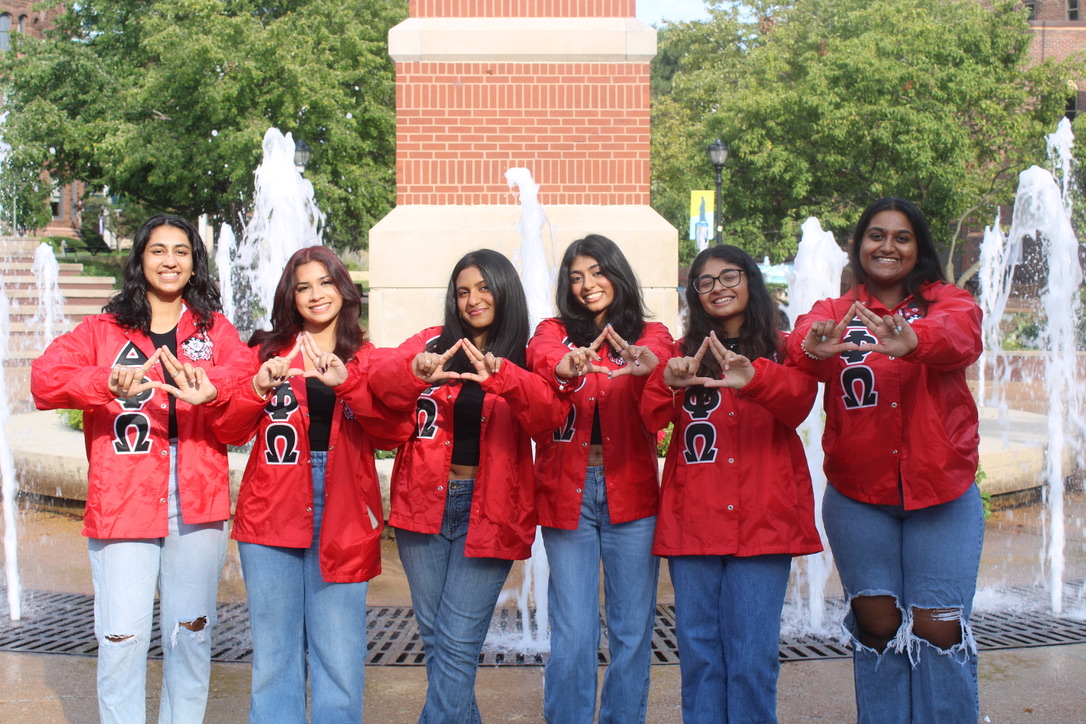 Members of SLU's Delta Phi Omega Sorority, Inc. Sorority members wearing matching jackets with the greek letters for Delta Phi Omega make a delta symbol with their hands while standing in front of the SLU clock tower.