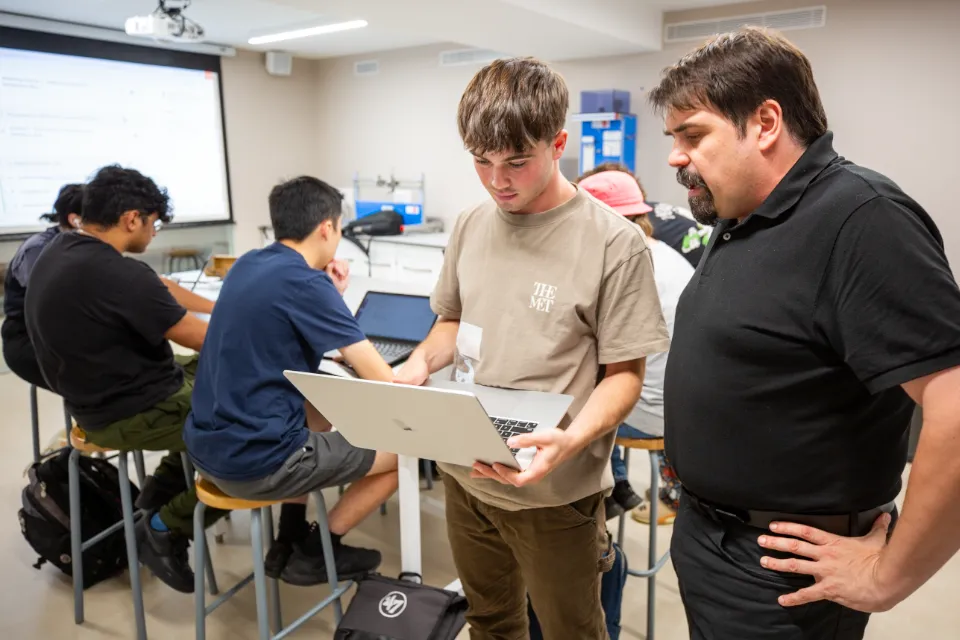 Academic Departments A student and a teacher look at a laptop while other students study seated behind them.
