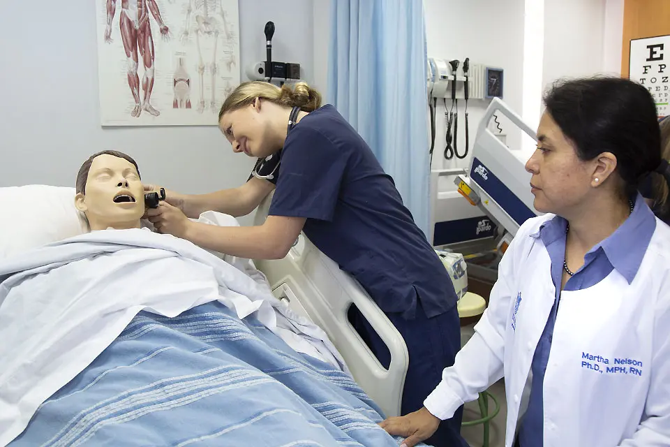 Health sciences at SLU-Madrid A student wearing medical scrubs checks the ear of a patient model on a hospital bed while an instructor observes.
