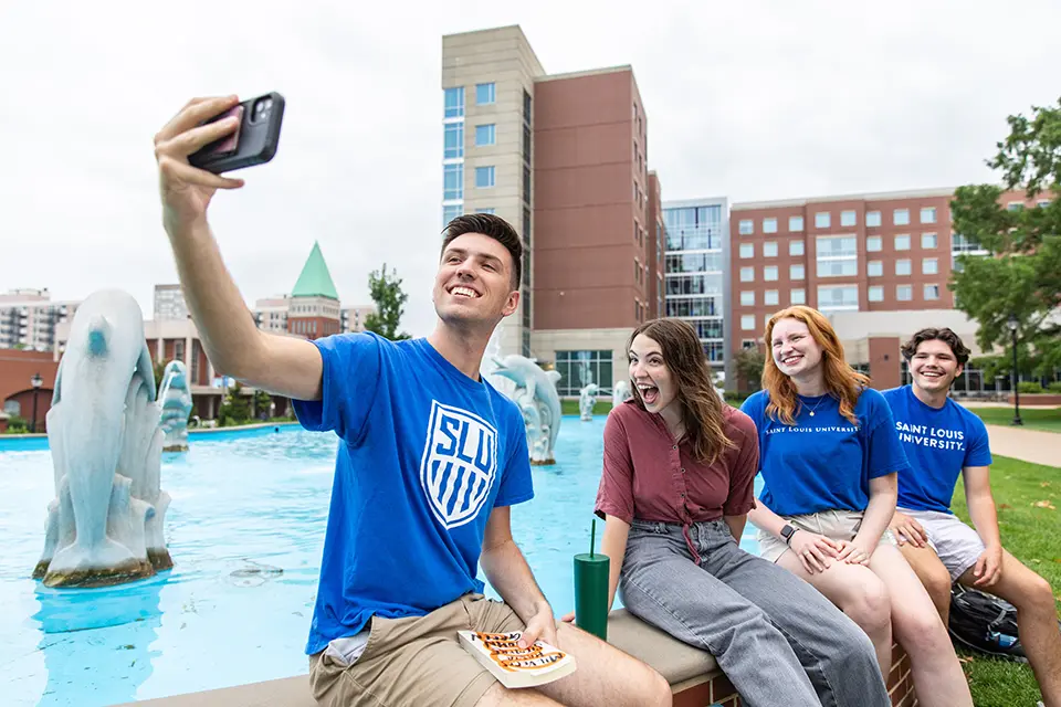 Madrid + St. Louis Majors A student takes a selfie while his friends look at the camera, sitting in front of a fountain.