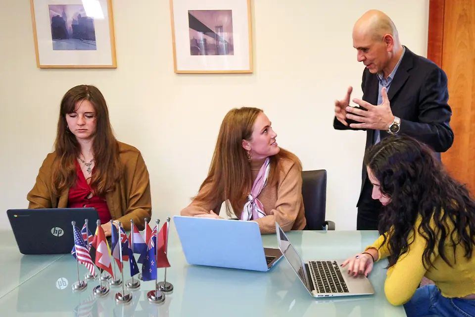 Three students with laptops, seated around a table with flags, attend to a teacher standing next to them.