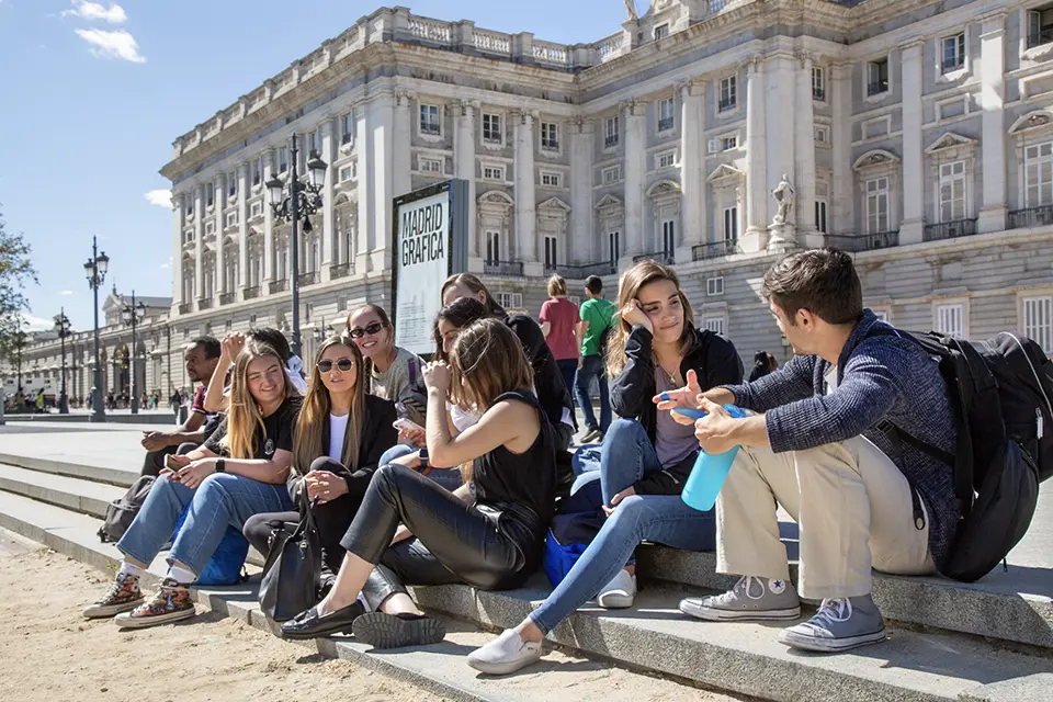 A group of students talk while sitting on steps in front of a large palace.