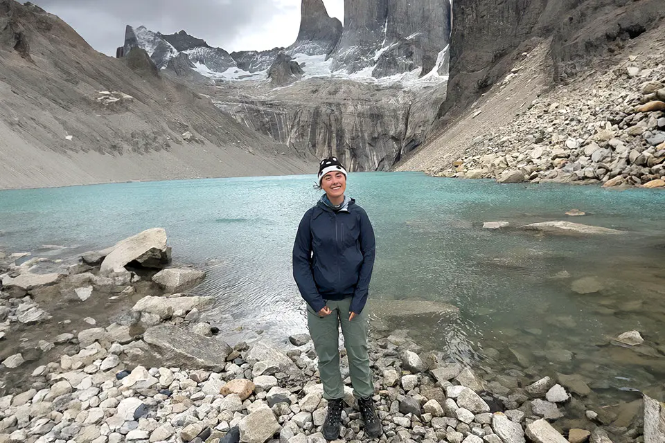 Quinn Riordan - Chile (Independent Non-SLU Study Abroad) A student wearing hiking gear stands on a rocky shoreline next to a body of water, surrounded by rocky cliffs and mountains.