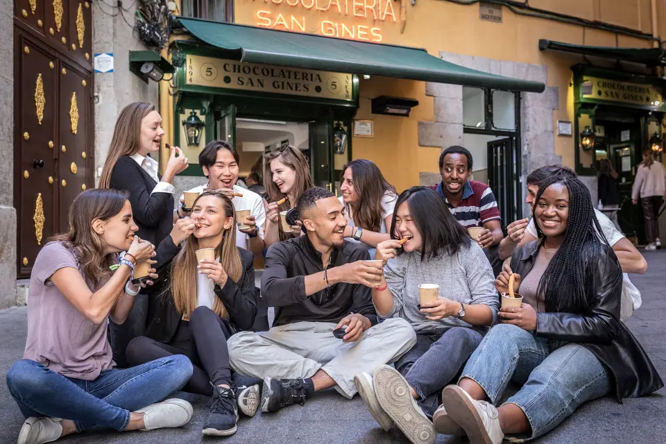 Students in Plaza Mayor in Madrid Students sit in front of a store called Chocolateria San Gines dunking pastries in cups of hot chocolate and eating them.