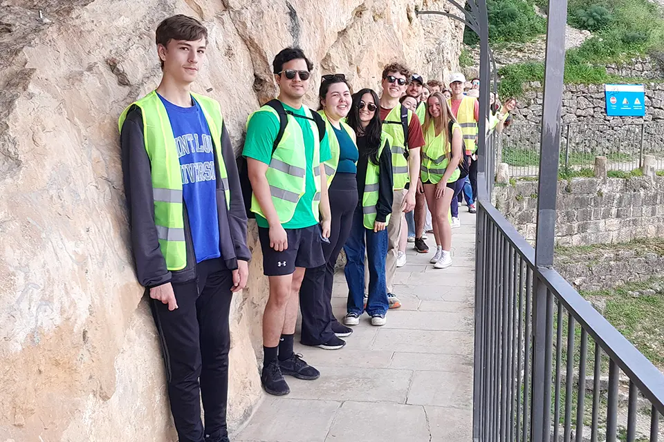 Geological Rocks Trip A group of students wearing reflective safety vests stands in a line on a walkway next to a rocky cliff side.