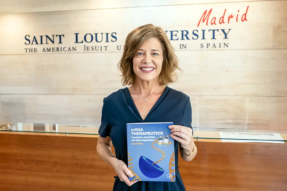 Anya Hillery, Ph.D., poses with her book "mRNA Therapeutics: Foundations, Innovations and Clinical Applications" in the San Ignacio Hall lobby. Woman holds book and poses for camera in front of reception desk with university logo on wall. Book title reads M R N A Therapeutics.