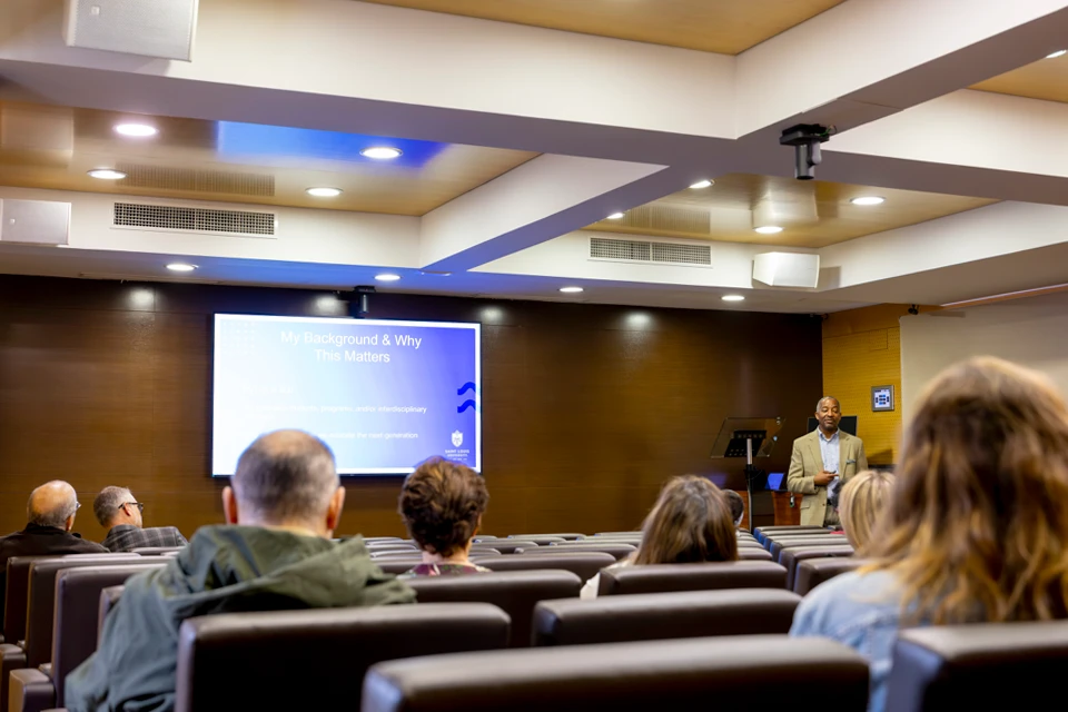 Triplett addresses audience at his presentation in San Ignacio Hall Auditorium. A man with a suit on looks at audience members who are sitting in a small auditorium with a screen that reads "My Background and Why This Matters."