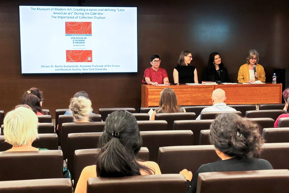 Four panelists sit at the front of a lecture hall while one speaks into a microphone. A screen displays a slide with the title: The Museum of Modern Art: Creating a Canon and Defining 'Latin American Art' During the Cold War — The Importance of Collection Displays