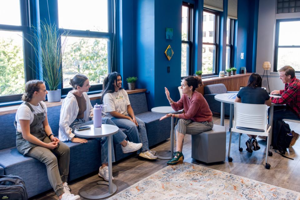 Pictured is Dr. Lauren Schwartz interacting with students in the new wellness space Students and a faculty member sit on chairs and benches in a room with windows and plants.