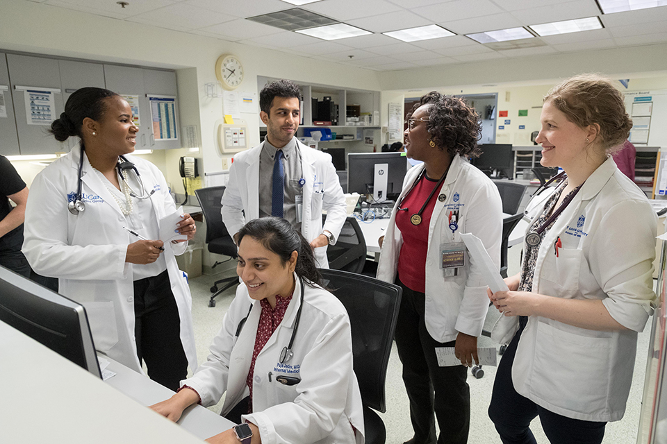 Diversity med students Four medical students stand behind a classmate who is seated and working at a computer. Students are smiling.