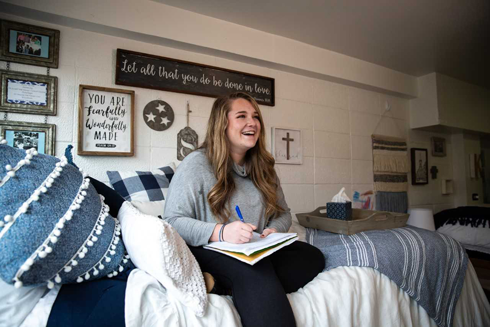 Savannah Smith, Class of 2022 Photo taken in Marguerite Hall A student sits on her bed in a dorm room, laughing while writing in a notebook.
