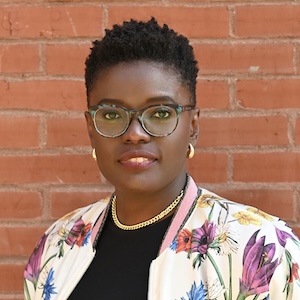 Bukky Gbadegesin A shoulder-length portrait of a woman with textured black hair, closely cropped on the sides, stands in front of a brick wall. She wears glasses with a blue tortoise-shell pattern, gold earrings, a gold chainlink necklace, and a black top with a floral-print jacket. Her smiles slightly gazes with a pleasant expression.