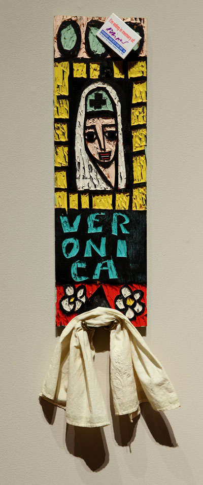 Adrian Kellard, Veronica towel rack, 1982. Latex on wood with hooks. Collection of Regina DeLuise, Baltimore. Photo by Jeffrey Vaughn An artwork by Adrian Kellard titled Veronica towel rack. A narrow rectangular carved piece of wood features the face of Veronica with her name below, various geometric patterns, and a hook with a towel hanging from it. The work is painted in black, yellow, teal, red and white.