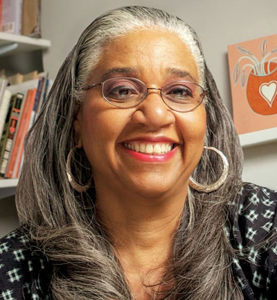 Leslie King-Hammond Curator and art historian Leslie King-Hammond, wearing large silver hoop earrings, sits in an office with books and a small painting in the background