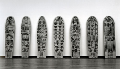 Maria Magdalena Campos-Pons, The Seven Powers Come By the Sea, 1992. Image courtesy of the artist. An artwork by Maria Magdalena Campos-Pons titled, The Seven Powers Come By the Sea. Seven ironing boards lean upright against a wall. Each bears a reproduction of a diagram of slaves packed into the hold of a slaving ship.