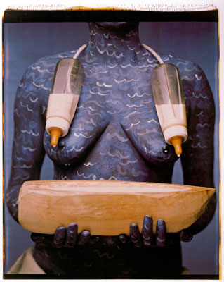 Maria Magdalena Campos-Pons, When I Am Not Here / Estoy Allá, 1994. Image courtesy of the artist. An artwork by Maria Magdalena Campos-Pons titled, When I Am Not Here / Estoy Allá. A photograph of the torse of a woman who is painted in blue with white waves, holding a stylized wooden boat. Two baby bottles half-filled with milk are connected by a cord behind her neck and suspended over her chest.