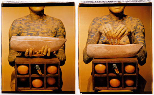 Maria Magdalena Campos-Pons, When I Am Not Here / Estoy Allá, 1994. Image courtesy of the artist. An artwork by Maria Magdalena Campos-Pons titled, When I Am Not Here / Estoy Allá. Two photographs of the torso of a women, her skin painted blue with white waves, kneeling behind a box with six cubbies containing various objects. In the left panel, she holds a stylized wooden boat. In the right panel, honey drips into the boat from her fingers.