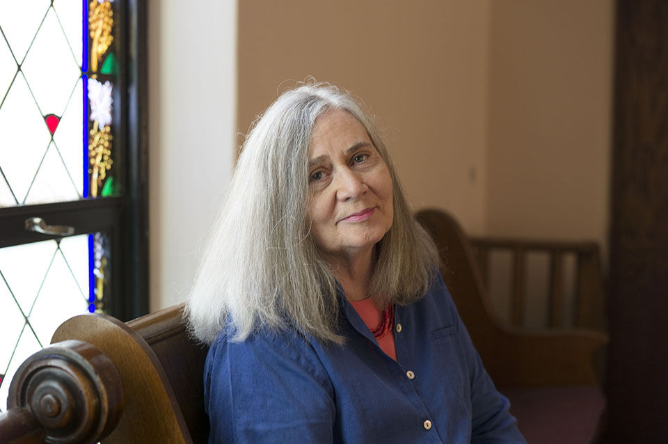  Author Marilynne Robinson, with long gray hair in a blue shirt, sits on a wooden bench next to a stained glass window.