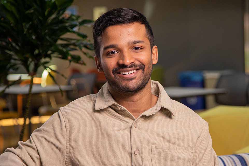 Akshay A man in a tan button up sits in an atrium with a plant in the background