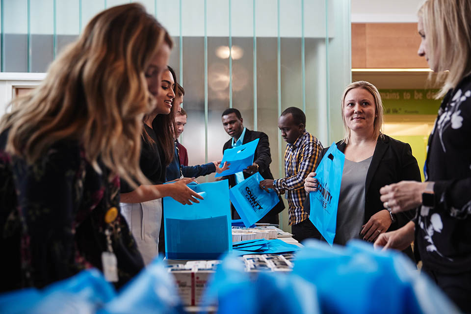 MPH Narcan Kit Packing People stand on both sides of a long table handling boxes and blue bags that say Saint Louis County